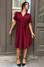 Woman wearing a burgundy dress standing in front of a building entrance. ANNICK