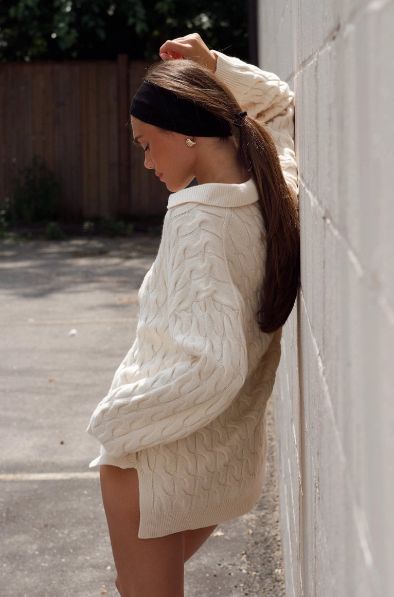 Woman wearing a white cable knit sweater leaning against a wall. Amanda Maria