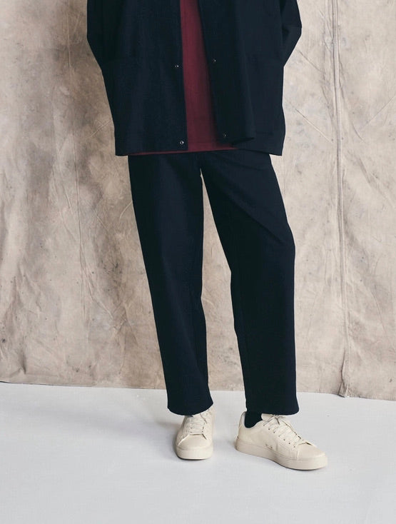 Man wearing a dark coat, maroon shirt, black pants, and white sneakers against a textured wall. Devlyn van Loon