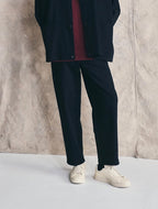 Man wearing a dark coat, maroon shirt, black pants, and white sneakers against a textured wall. Devlyn van Loon