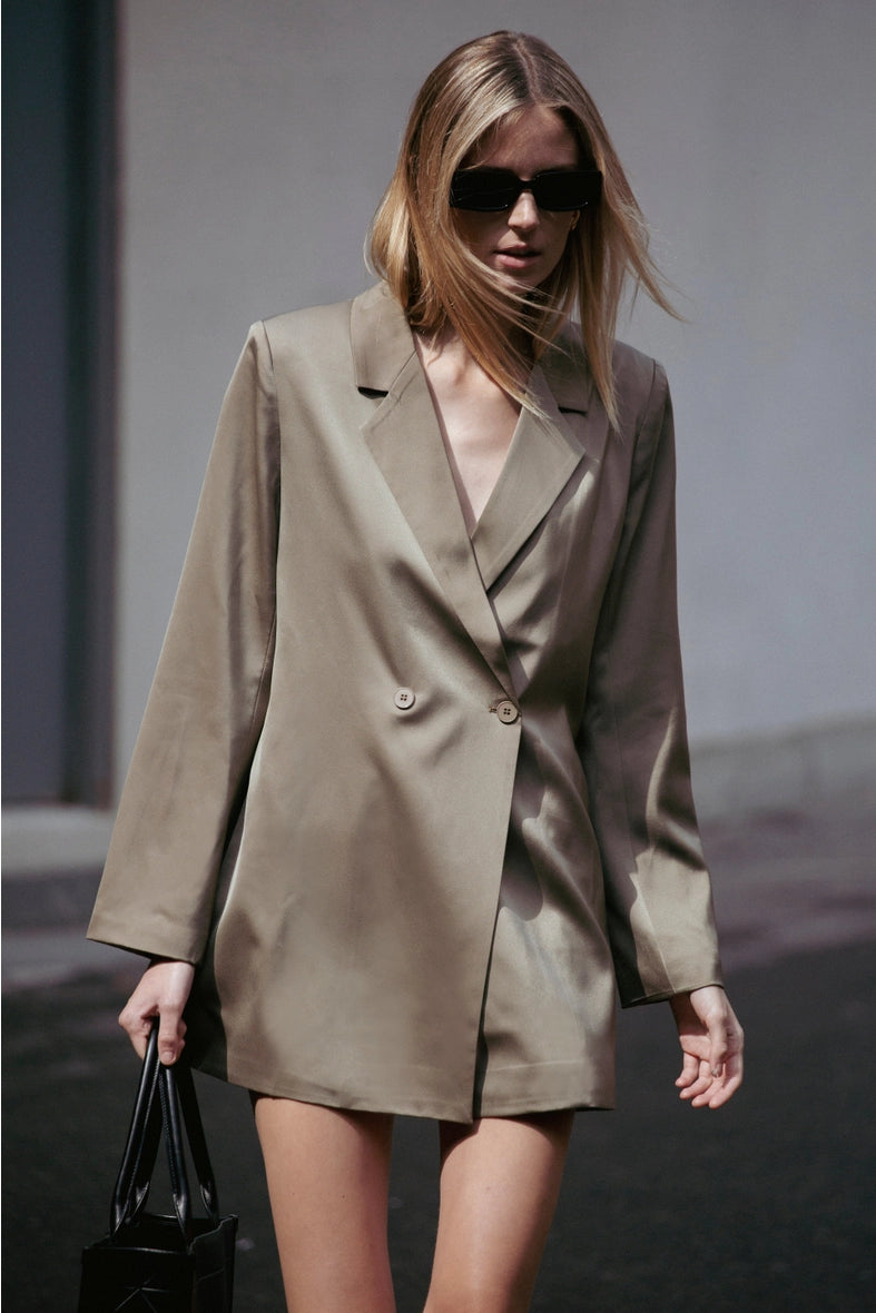 Woman wearing a beige trench coat and sunglasses, holding a black handbag.  Amanda Maria