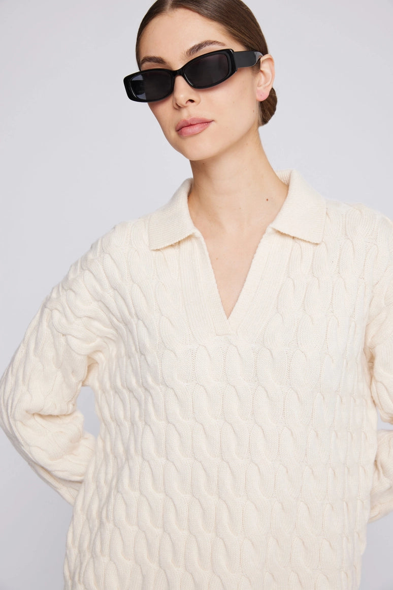 Women wearing a cream cable knit sweater and black sunglasses against a light gray background Amanda Maria