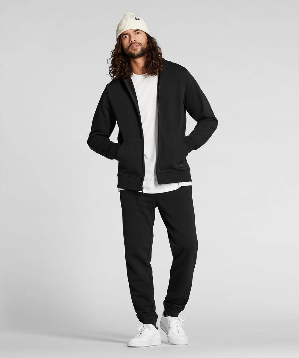 Man wearing a black hoodie, black pants, white shirt, white beanie, and white sneakers on a light gray background RYU