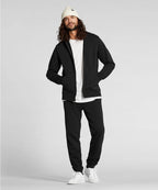 Man wearing a black hoodie, black pants, white shirt, white beanie, and white sneakers on a light gray background RYU