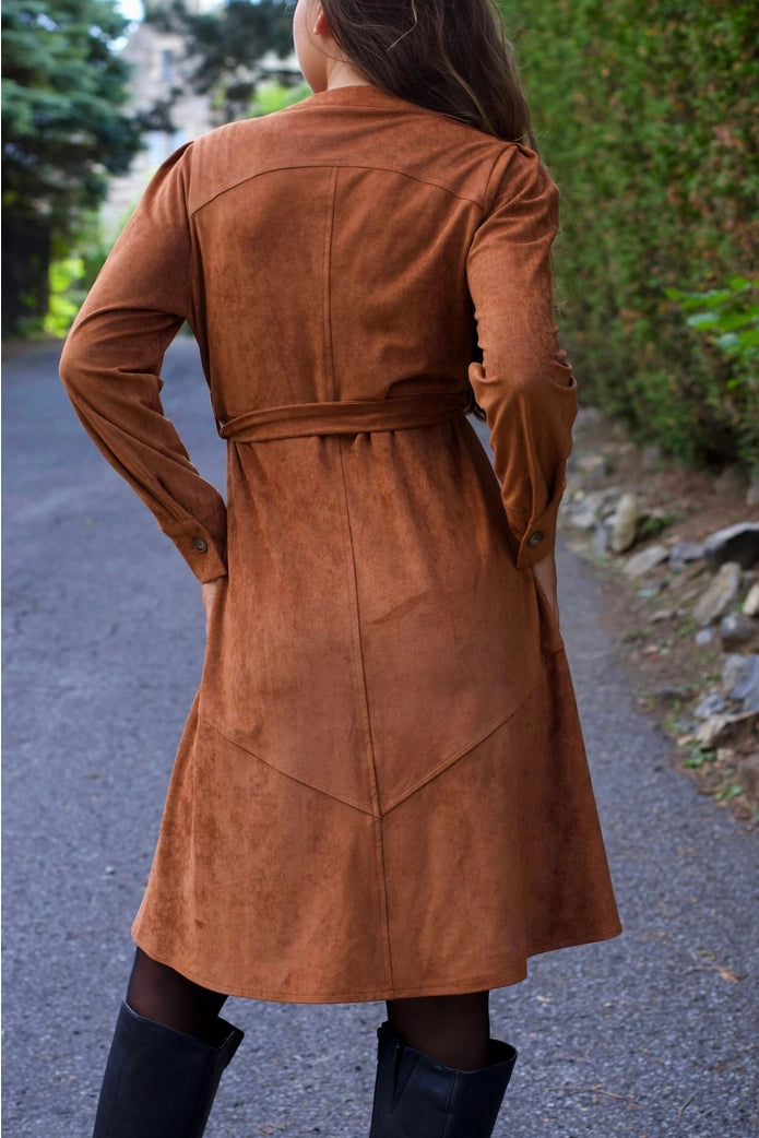 Women wearing a brown suede coat walking on a path with greenery. ANNICK