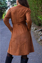 Women wearing a brown suede coat walking on a path with greenery. ANNICK