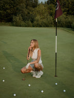 Woman on a golf course preparing to putt with a flag and golf balls in the background in a grey BOV skort