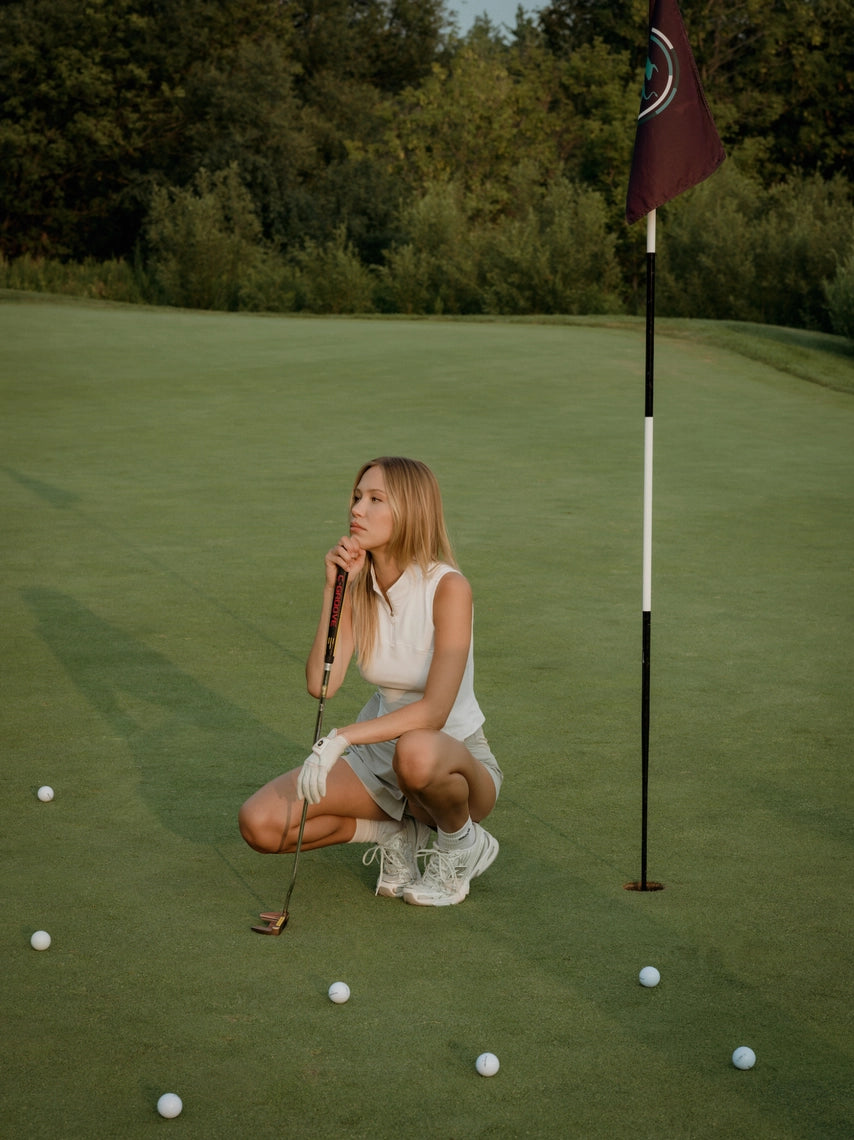 Woman on a golf course preparing to putt with a flag and golf balls in the background in a grey BOV skort