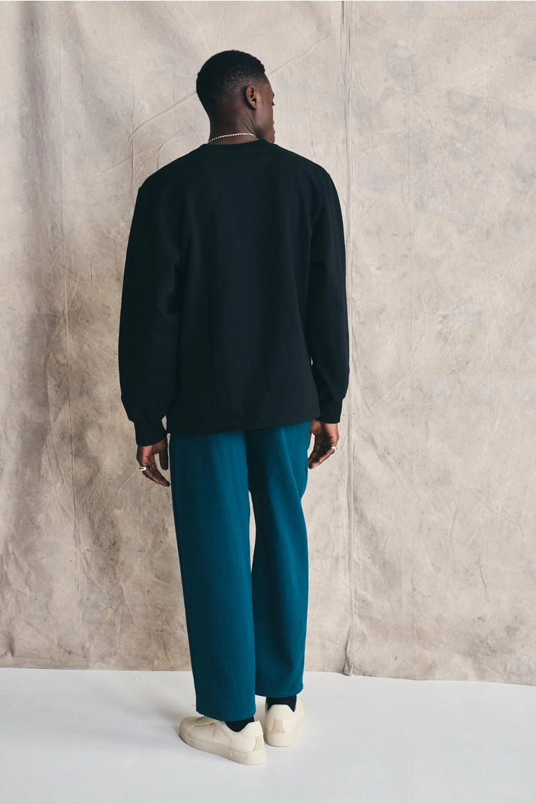 Man wearing a black sweater and teal pants standing against a beige wall. Devlyn van Loon