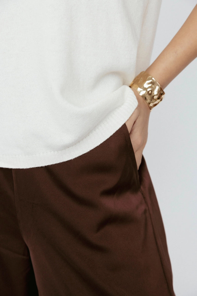 Person wearing a white top and brown pants with a gold bracelet on a plain background Amanda Maria