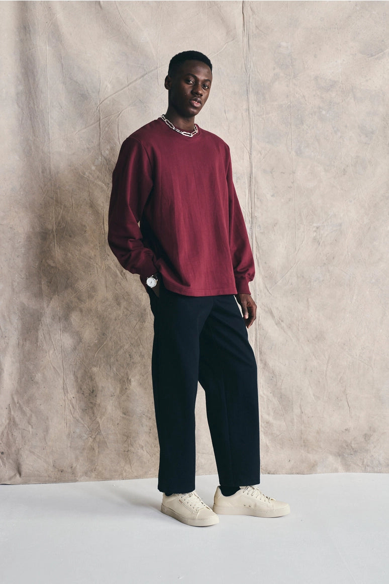 Man wearing a burgundy sweater, navy pants, and white shoes against a beige wall. Devlyn van Loon