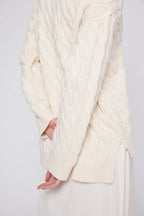 Close-up of a person wearing a cream-colored cable knit sweater on a plain background Amanda Maria