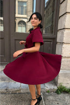 Woman in a burgundy dress standing outdoors next to a building. ANNICK