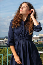 Woman in a navy blue dress standing outdoors with a cityscape in the background ANNICK