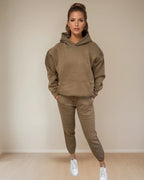 Woman wearing a brown hoodie and pants set with white sneakers against a beige background PRIV
