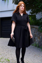 Woman wearing a black dress standing outdoors on a paved path. ANNICK