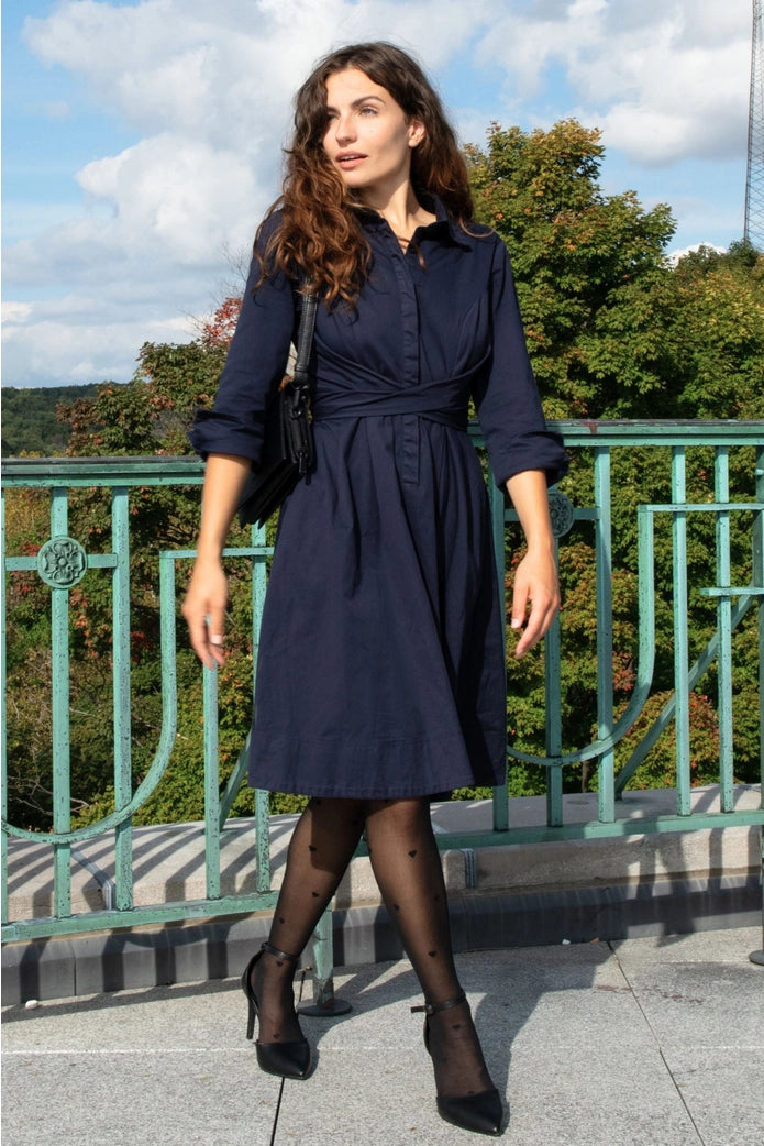 Woman in a navy dress standing outdoors with greenery and a blue sky in the background ANNICK