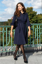 Woman in a navy dress standing outdoors with greenery and a blue sky in the background ANNICK