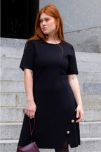 Woman wearing a black dress with gold buttons on gray steps ANNICK