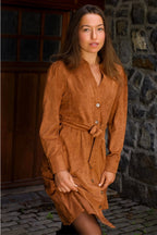 Woman wearing a brown dress standing against a stone wall. ANNICK