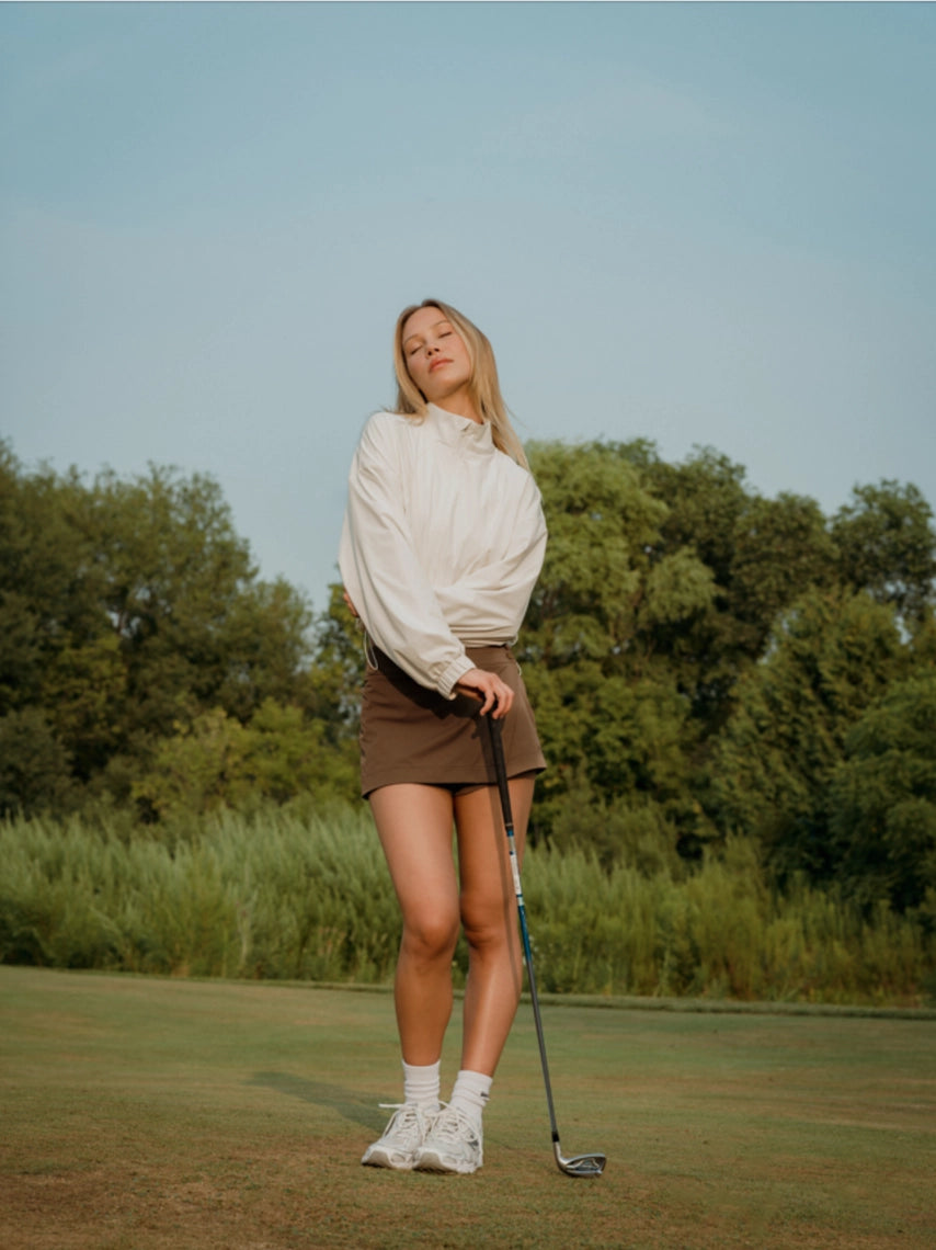 Woman holding a golf club on a golf course with trees in the background in a brown BOV skort