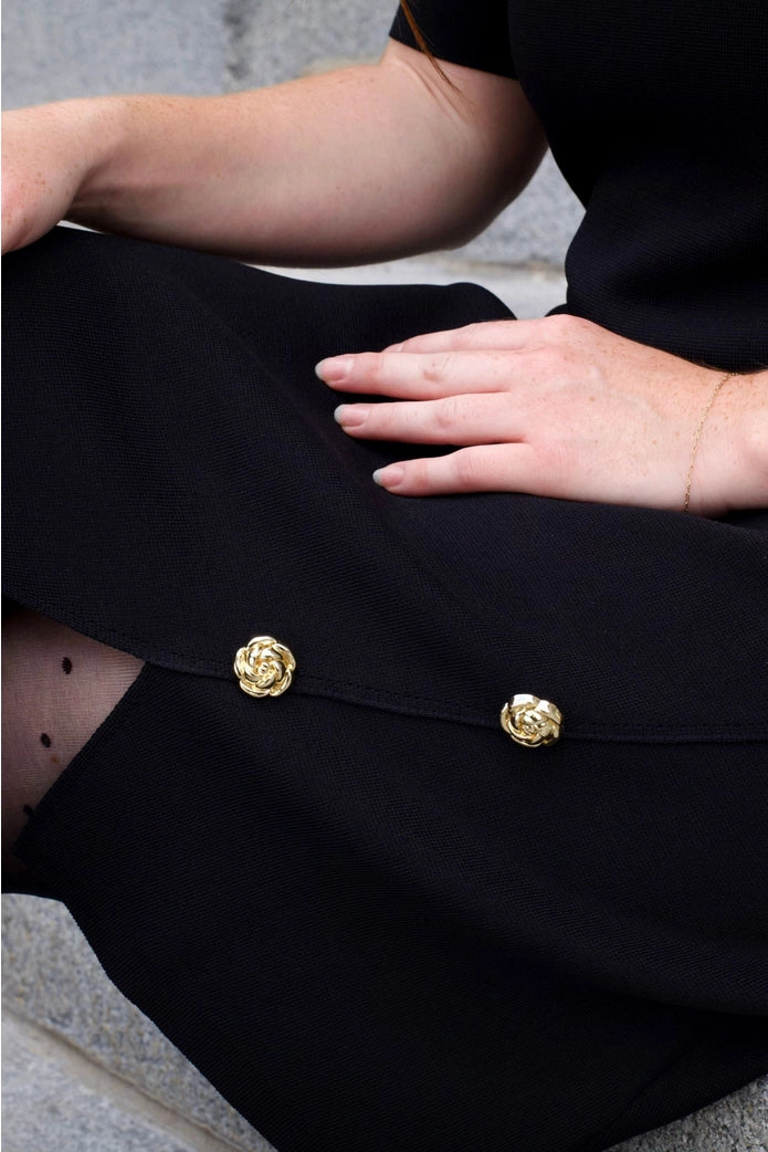 Women wearing a black outfit with gold buttons, sitting on a gray surface. ANNICK