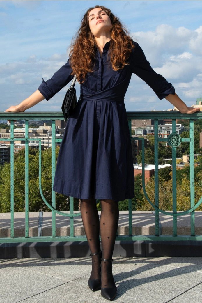 Woman in a navy dress standing on a rooftop with a cityscape in the background ANNICK