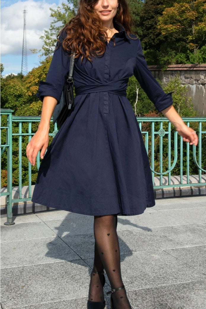 Woman wearing a navy blue dress standing outdoors with greenery in the background ANNICK