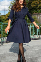 Woman wearing a navy blue dress standing outdoors with greenery in the background ANNICK