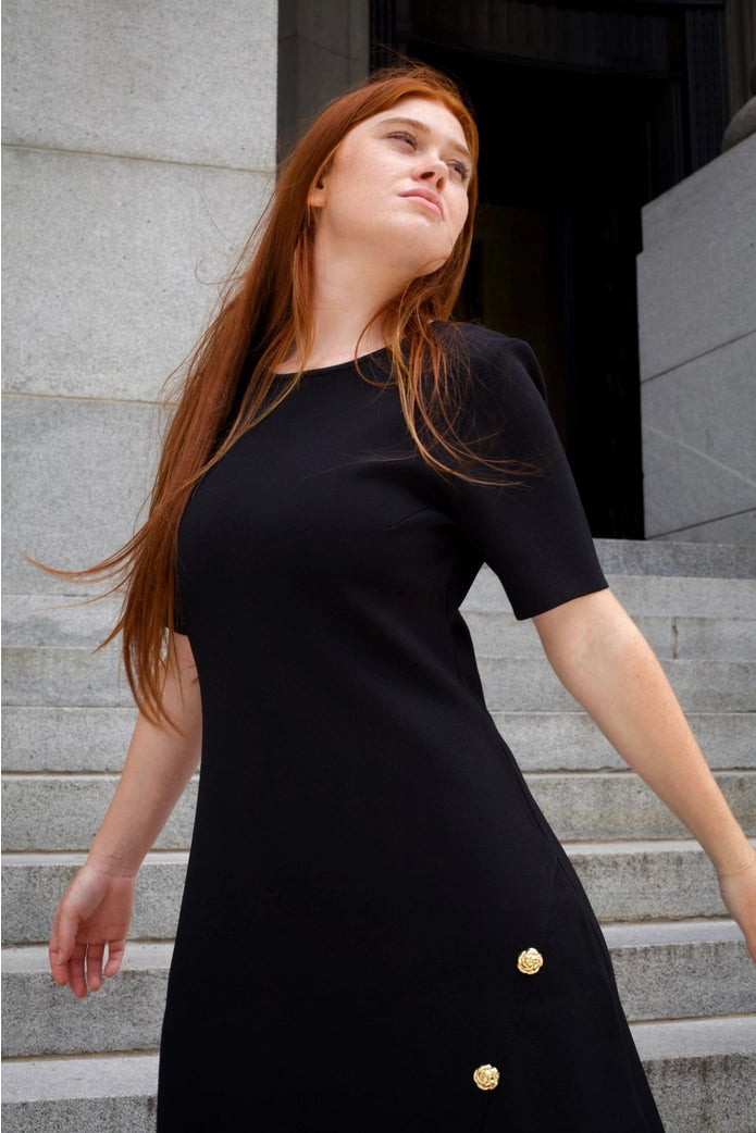 Woman in a black dress with gold buttons standing on steps. ANNICK