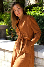 Woman wearing a brown dress standing outdoors with greenery in the background ANNICK
