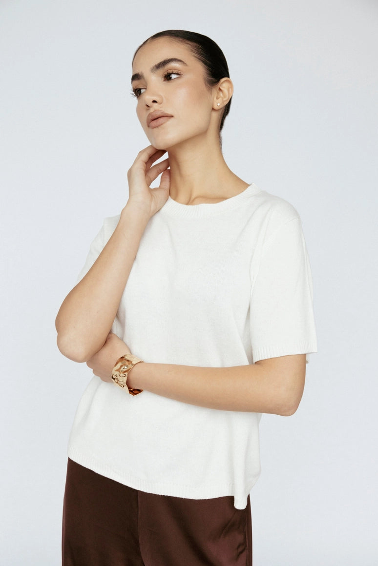 Woman wearing a white top and brown pants against a plain background Amanda Maria