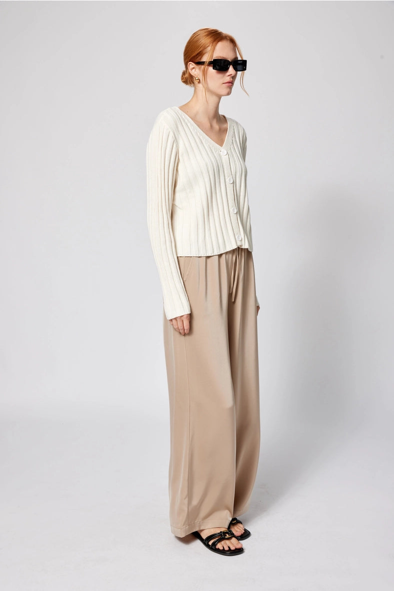 Woman wearing a white top and beige pants against a plain background Amanda Maria