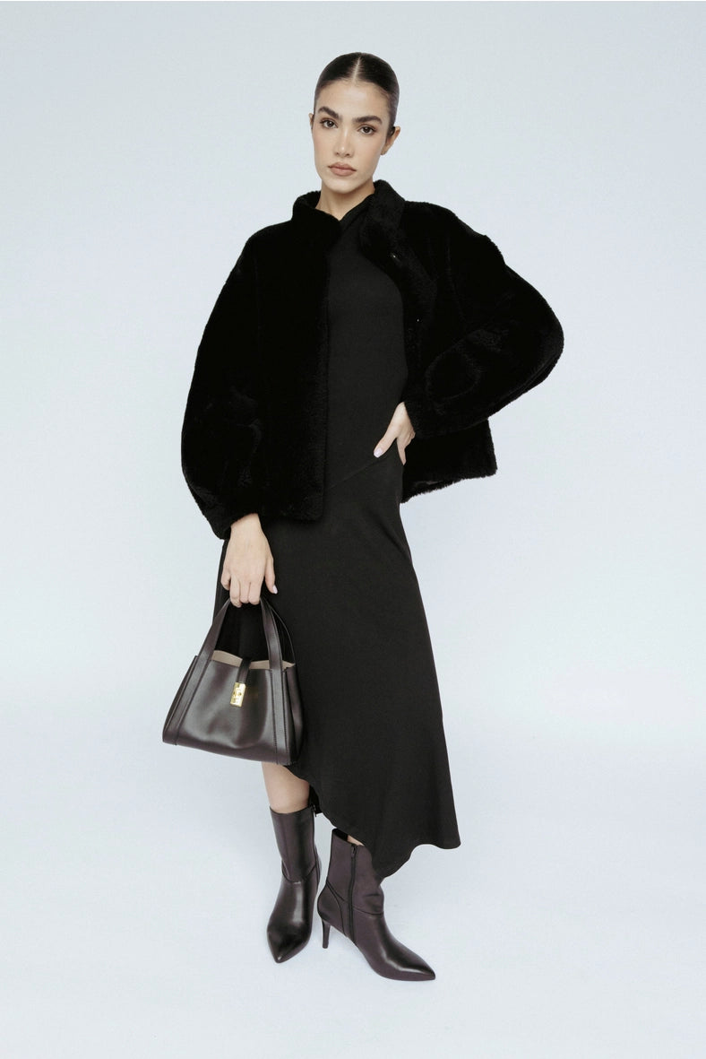 Woman wearing a black coat, black dress, and brown boots holding a black handbag on a white background Amanda Maria