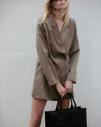 Woman wearing a brown blazer dress holding a black bag against a plain background Amanda Maria
