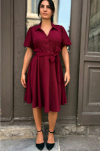 Woman wearing a burgundy dress standing in front of a door. ANNICK