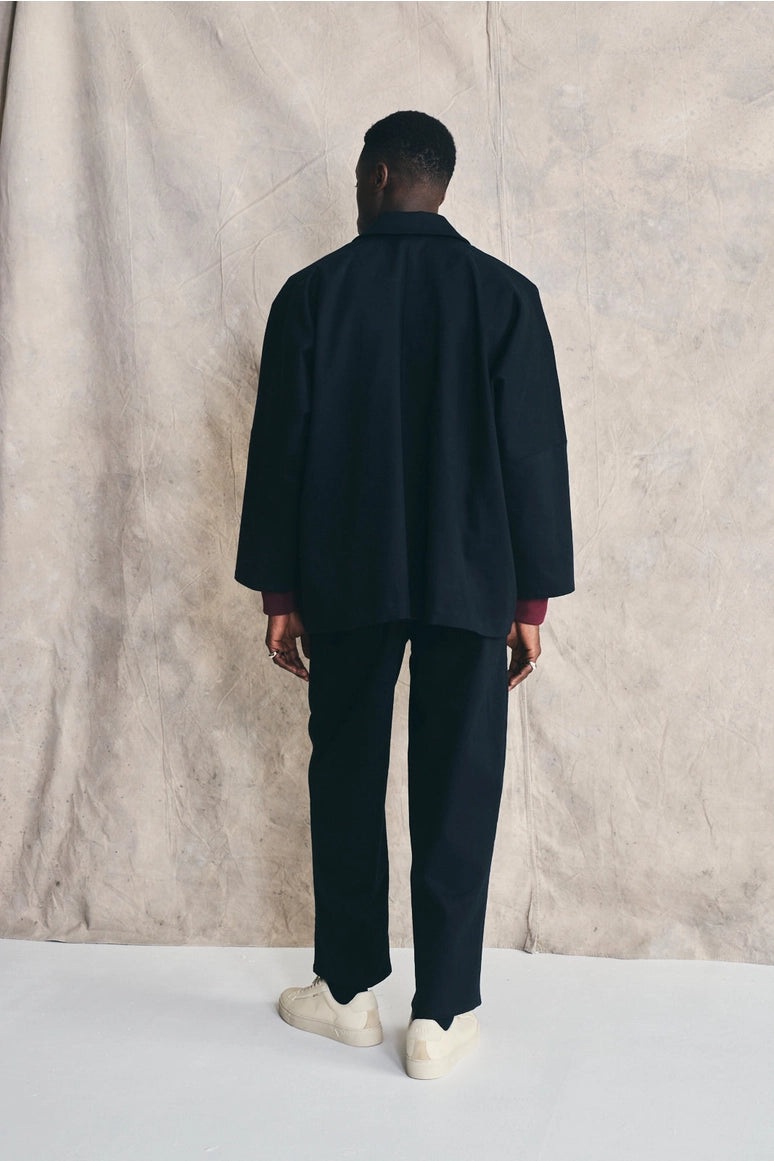 Man wearing a dark coat and pants standing against a textured beige wall. Devlyn van Loon