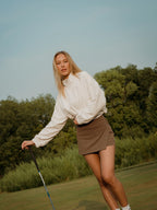 Woman on a golf course holding a golf club with trees in the background in a brown BOV skort