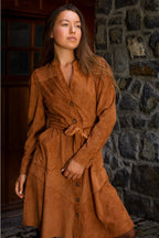 Woman wearing a brown suede dress standing against a stone wall. ANNICK