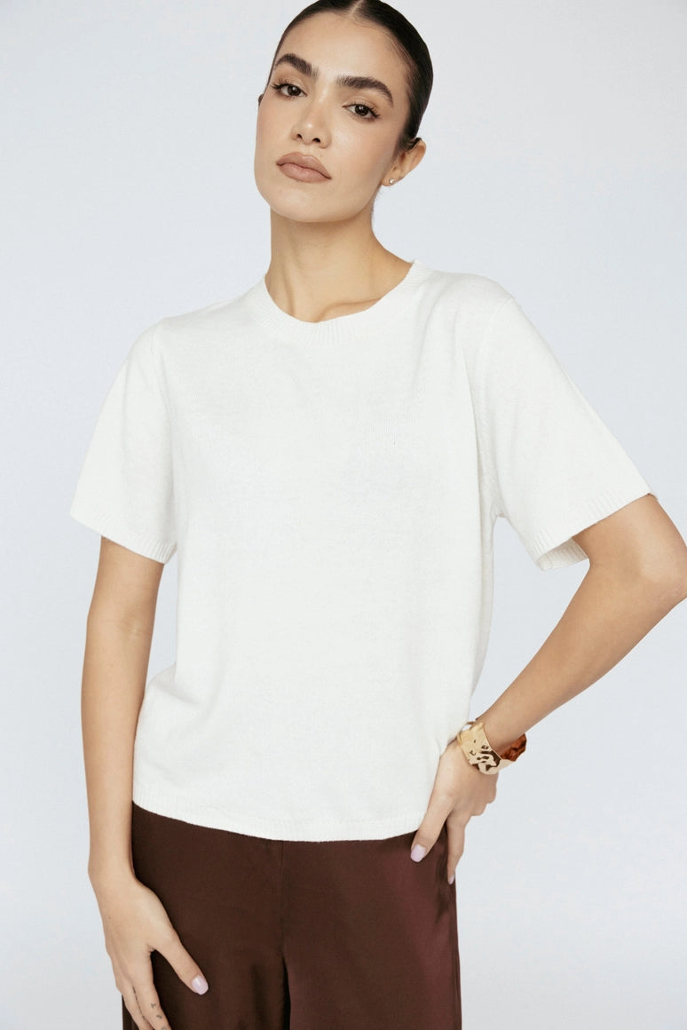 Woman wearing a white t-shirt and brown pants against a plain background Amanda Maria