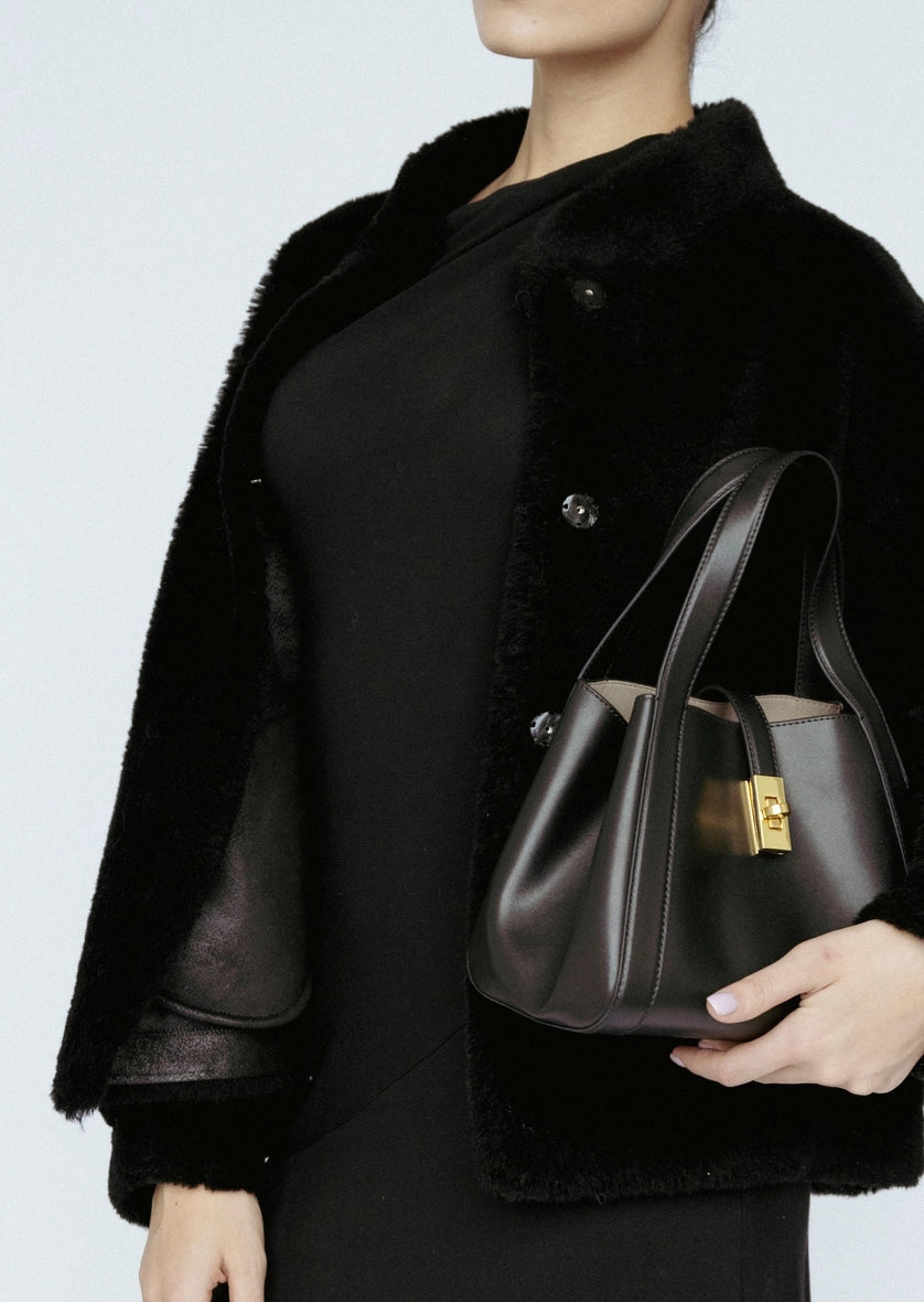 Woman wearing a black coat holding a dark handbag against a light background Amanda Maria