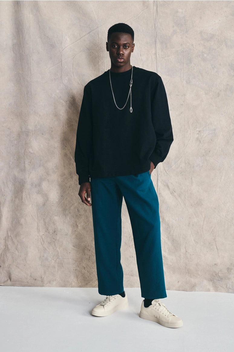 Man wearing a black sweater, teal pants, and white sneakers against a plain background Devlyn van Loon