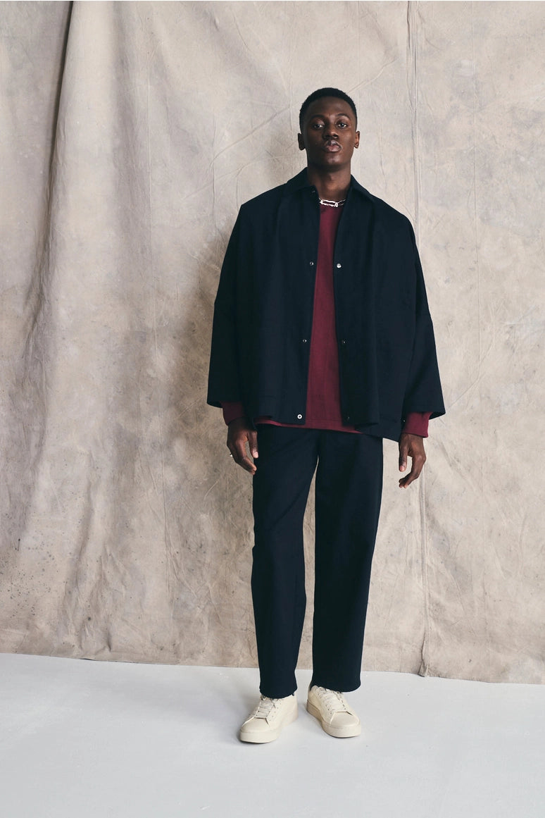 Man wearing a dark jacket, red shirt, and dark pants against a beige curtain background Devlyn van Loon 
