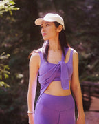 Woman in Rose Boreal purple athletic wear and cap standing outdoors with greenery in the background