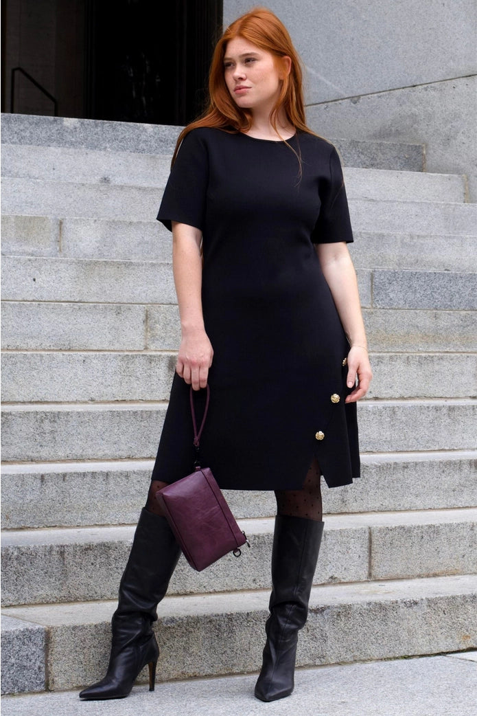 Woman in a black dress with gold buttons standing on stone steps. ANNICK