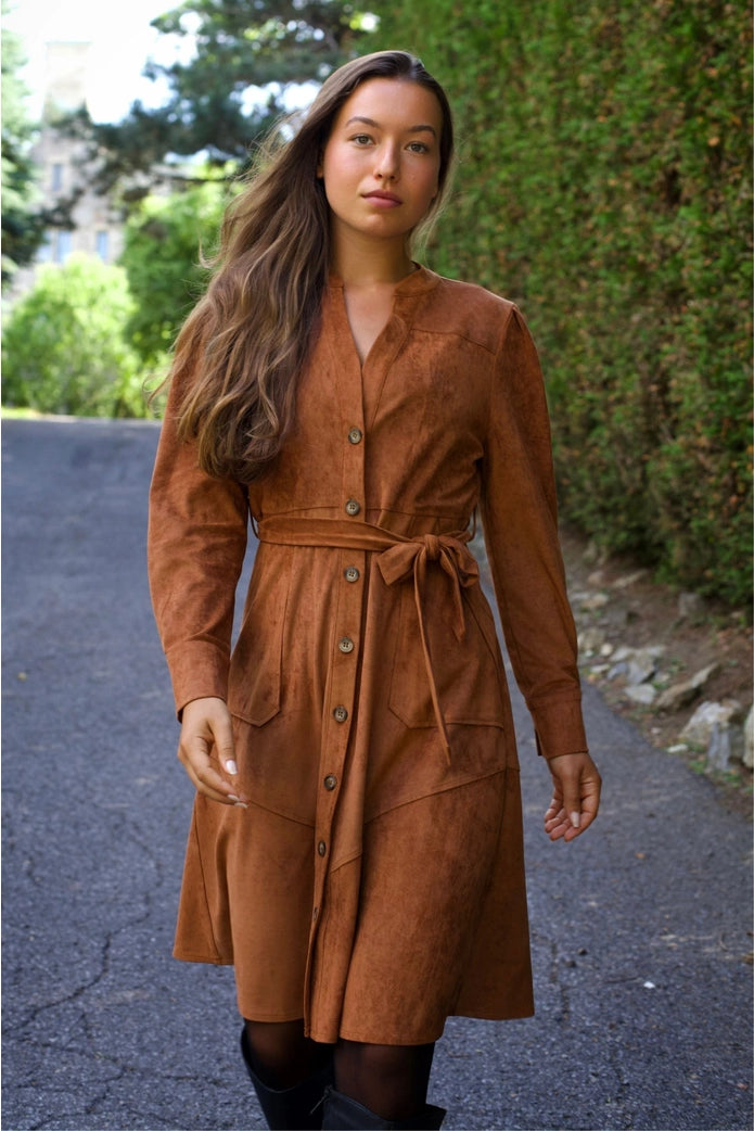 Woman wearing a brown suede dress standing on a road with greenery in the background ANNICK
