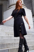 Woman in a black dress with gold buttons standing on stone steps. ANNICK