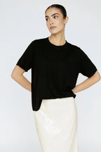 Woman wearing a black top and white pants against a light gray background Amanda Maria