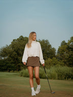 Woman walking on a golf course holding a golf club with trees in the background in a brown BOV skort