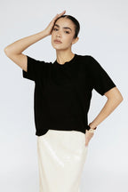 Woman wearing a black top and white pants against a light gray background Amanda Maria